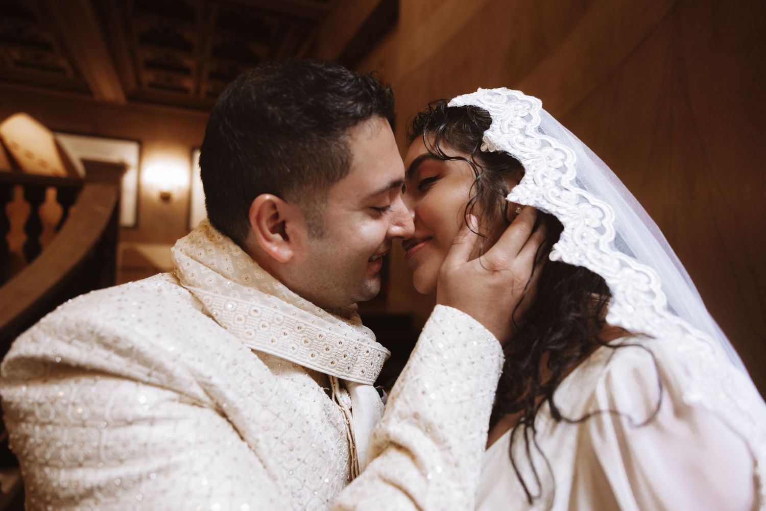 Groom in cream sherwani and bride in white veil share a tender kiss on staircase.