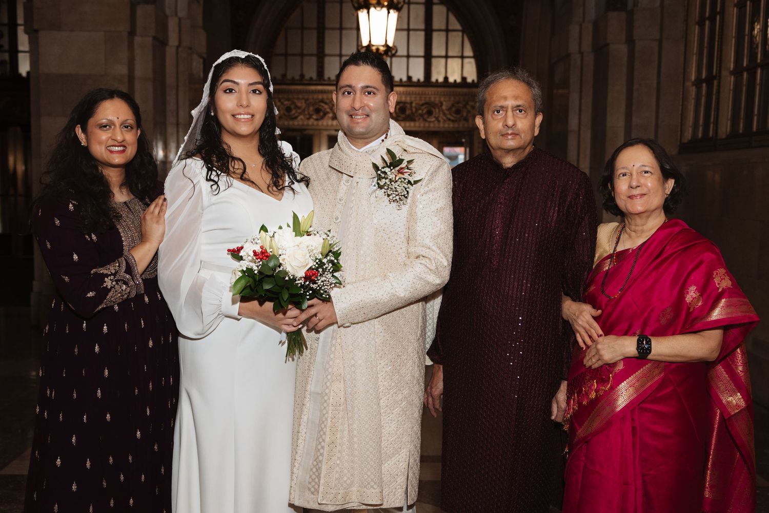 Newlyweds in traditional wedding attire pose with four family members in elegant Indian outfits at wedding ceremony.