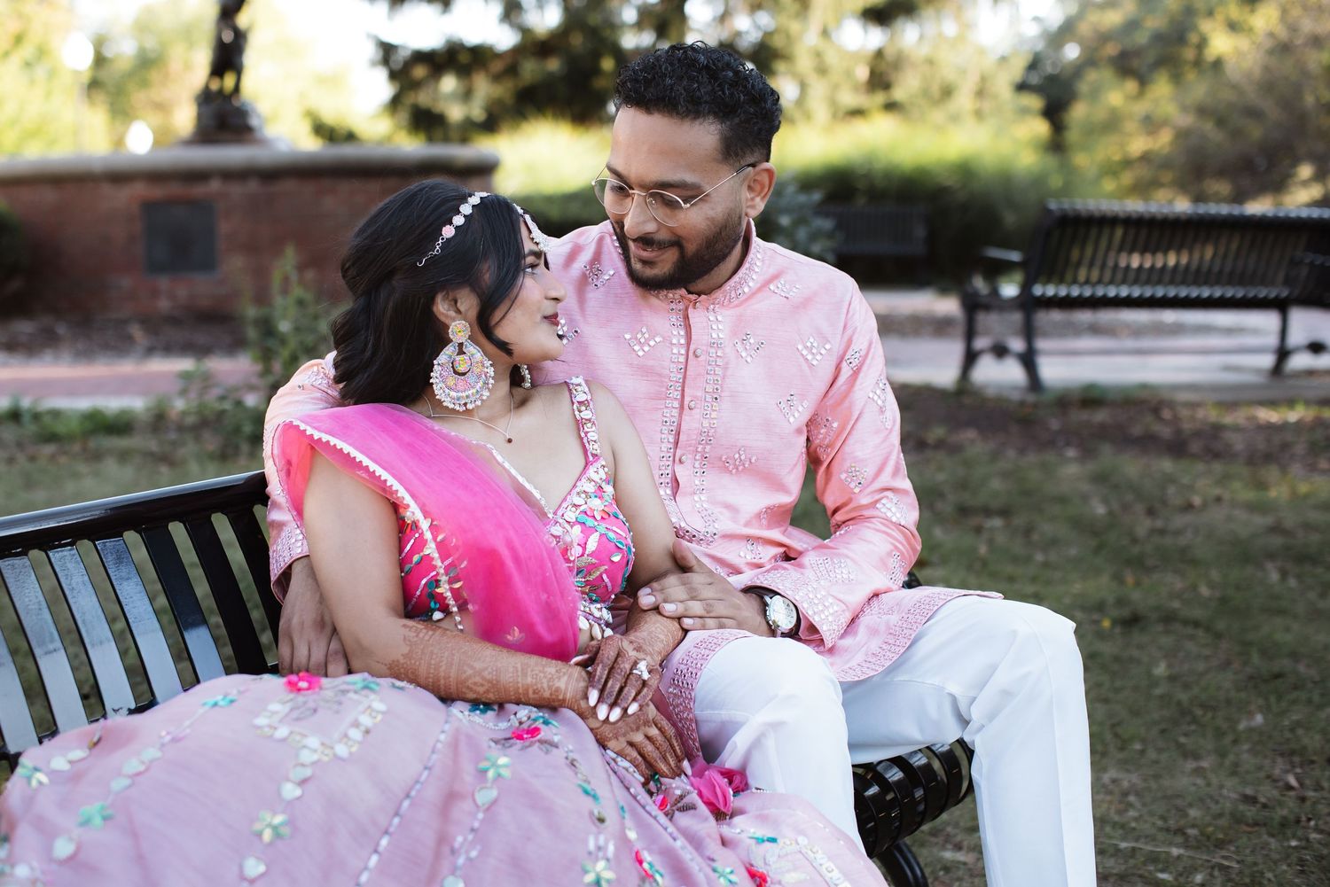 Indian bride and groom laughing together on a park bench in pink traditional attire.