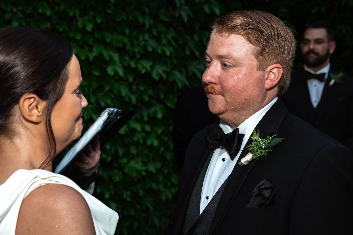 Groom in black tuxedo with boutonniere looks at bride during outdoor wedding ceremony.