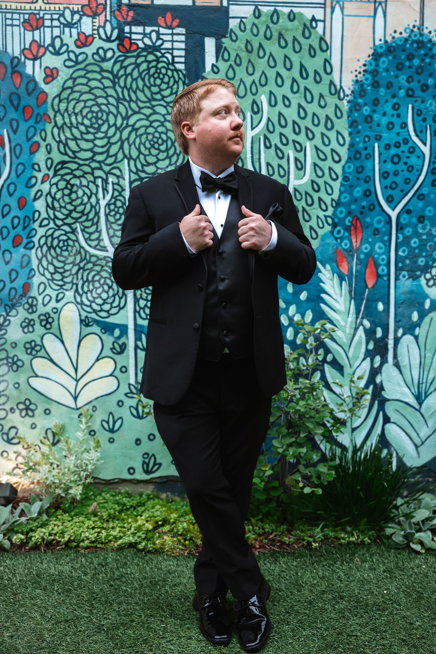 Stylish groom in black tuxedo adjusts lapels while posing in front of a colorful botanical mural.