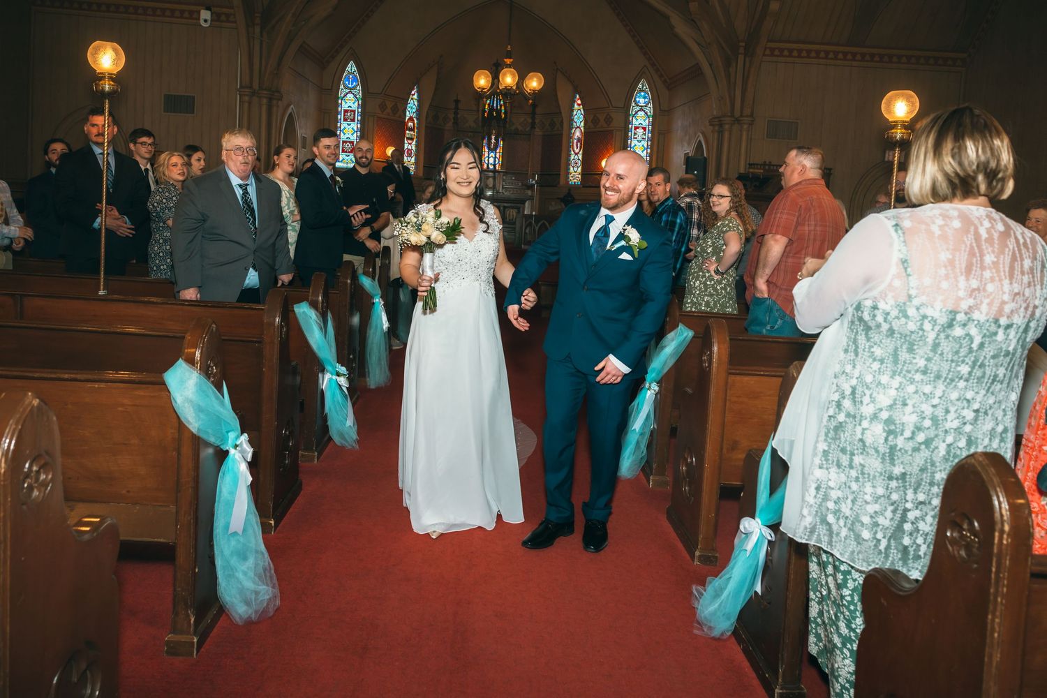 Newlyweds walk joyfully down the aisle of a decorated church with teal ribbon pew bows after their ceremony.