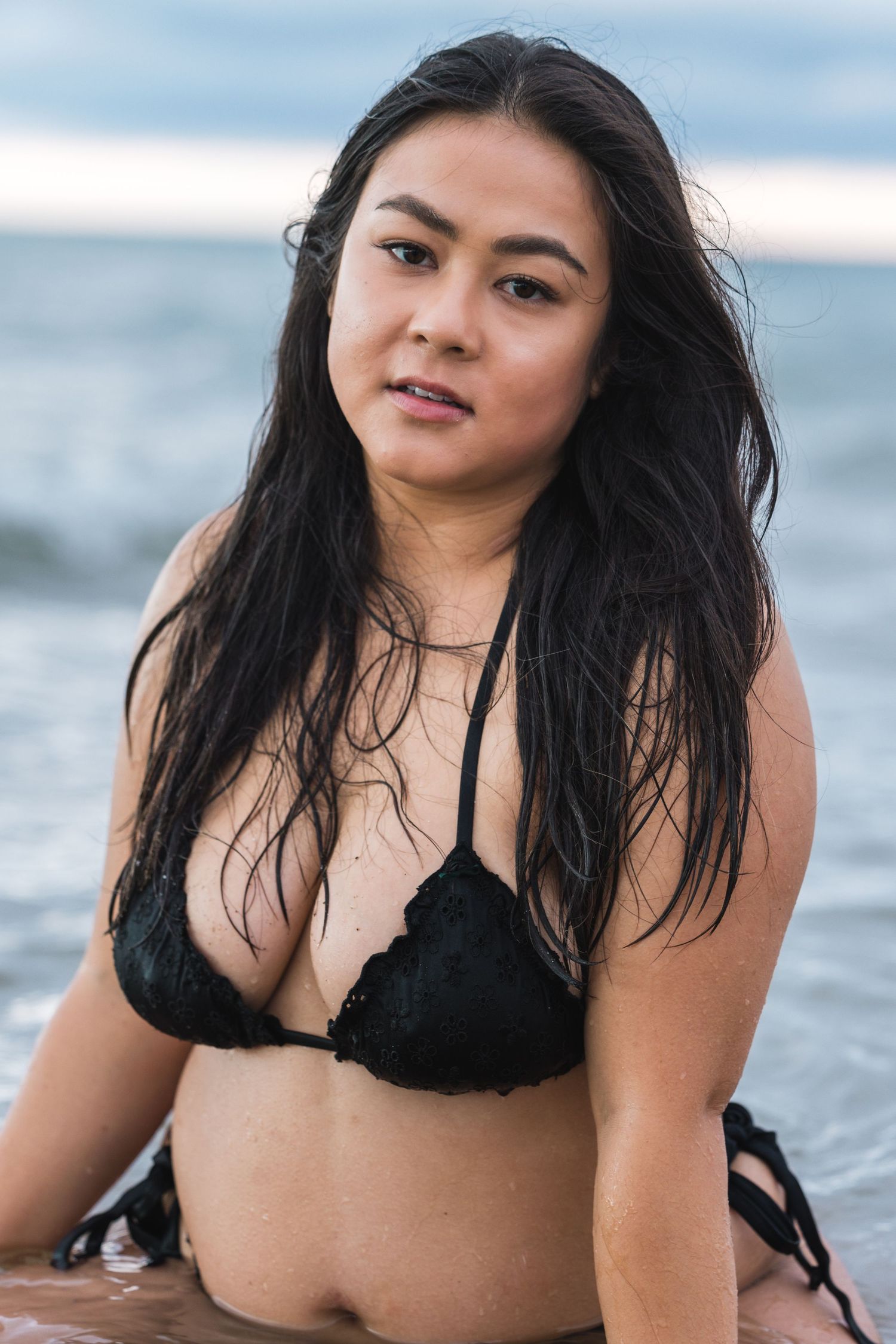 Woman in black bikini posing in shallow ocean water with wet dark hair on overcast day.