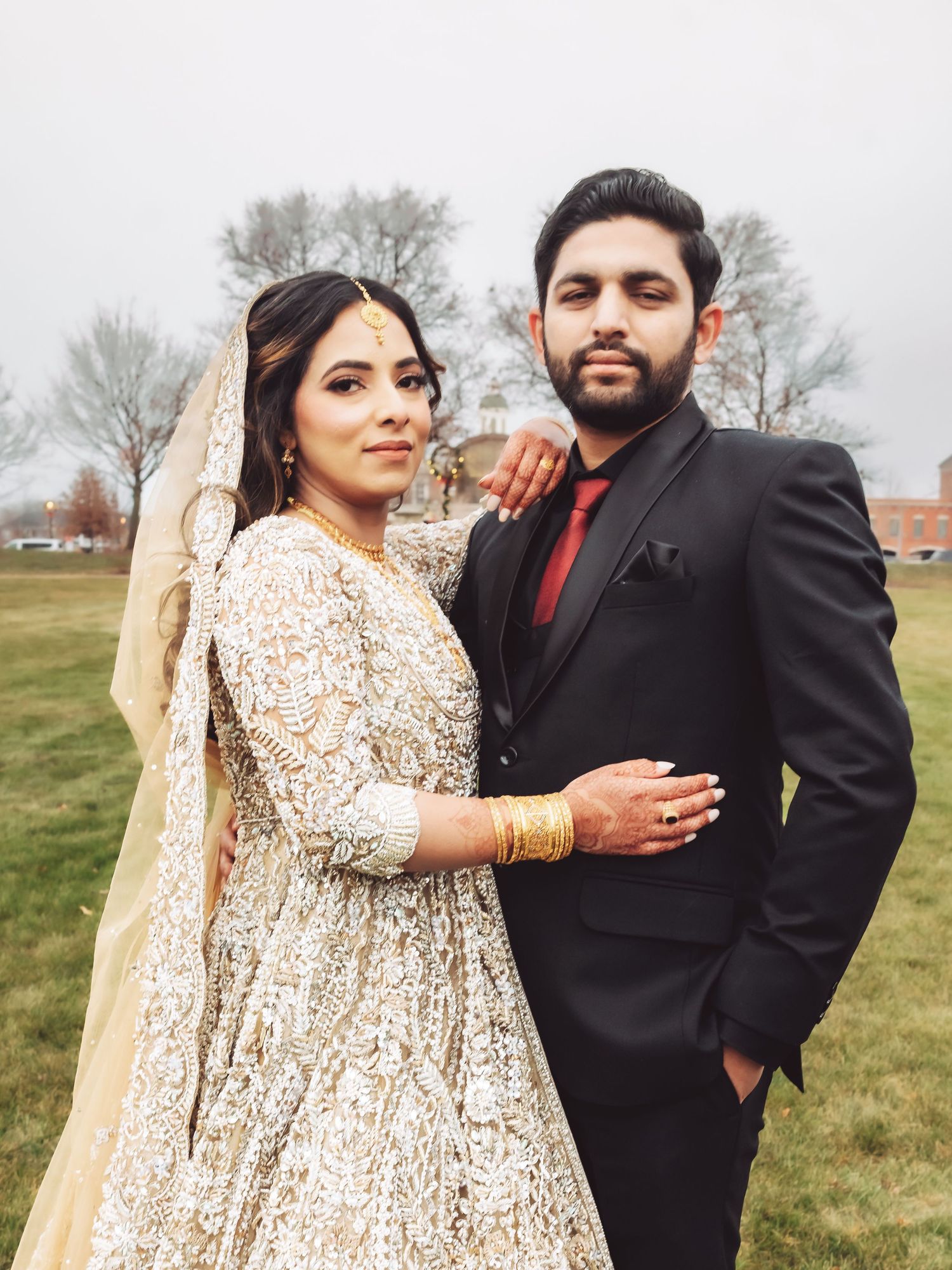 South Asian wedding couple posing outdoors, bride in gold lehenga, groom in black suit.