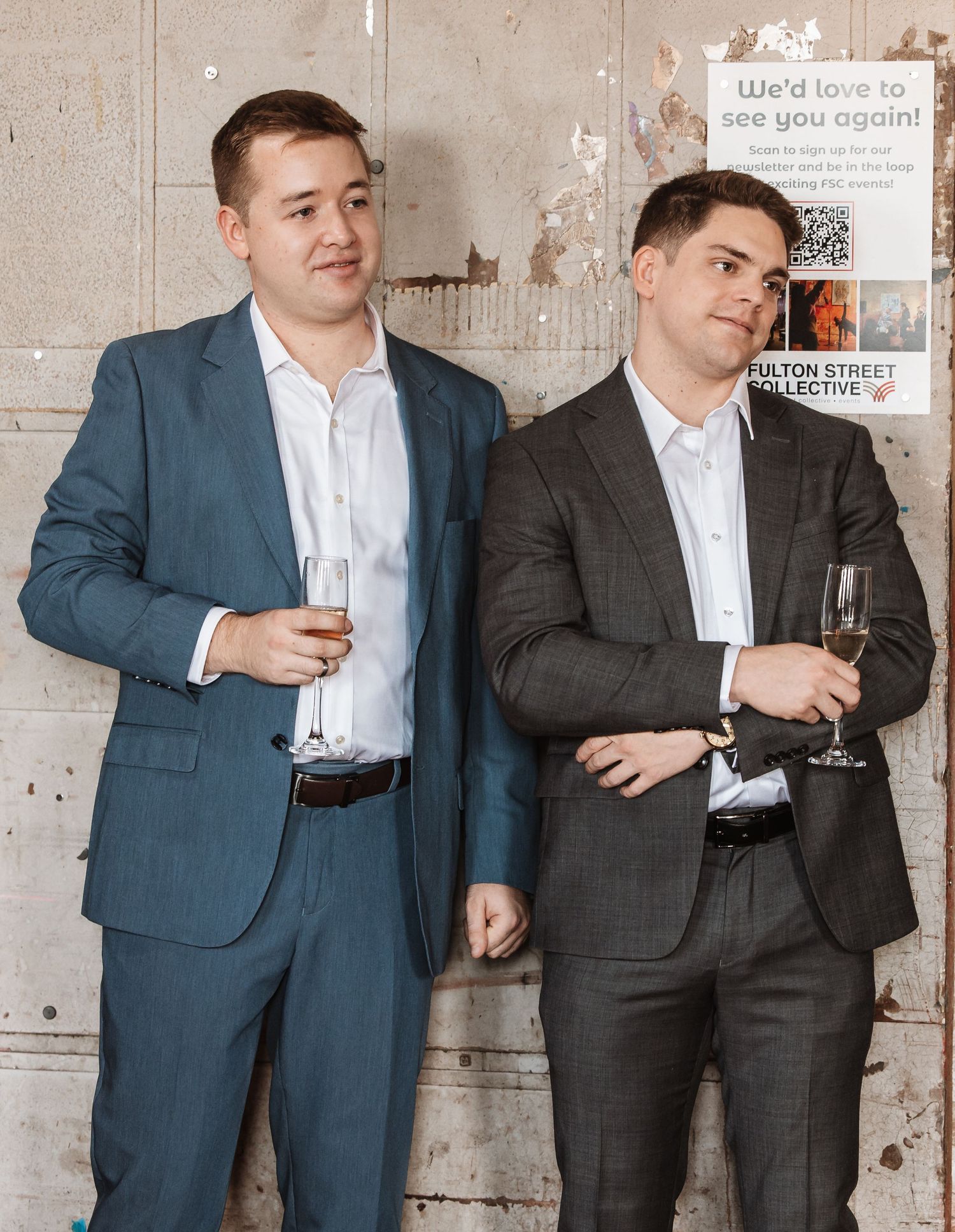 Two men in suits holding champagne glasses at a formal event near a brick wall.