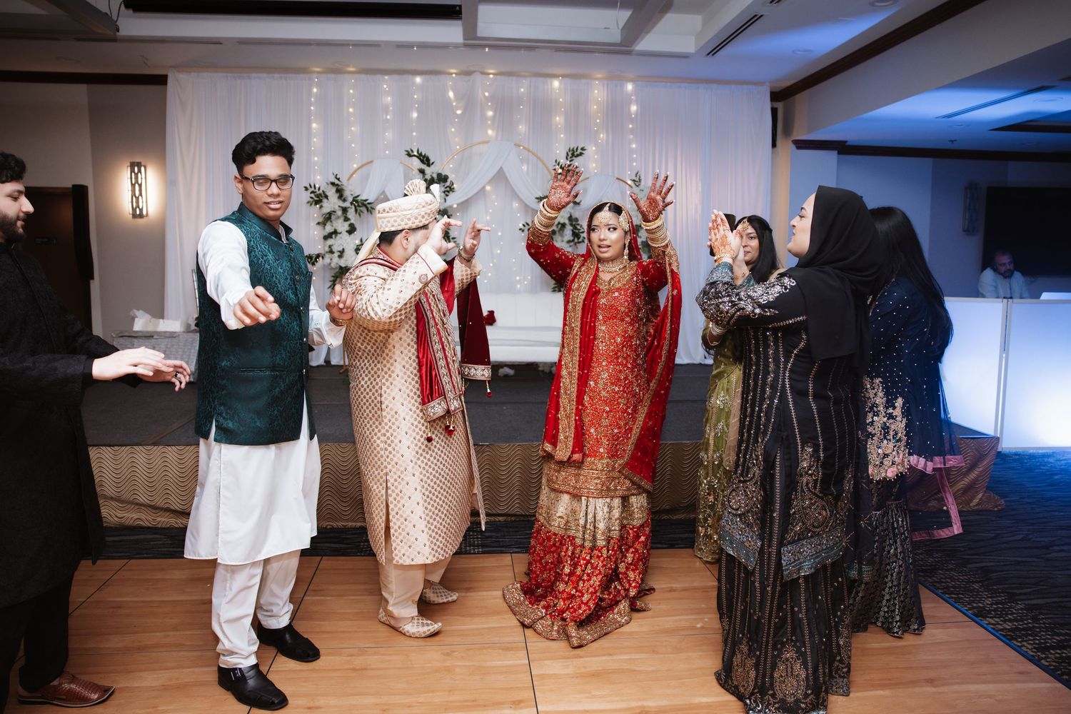 South Asian wedding couple in traditional attire celebrate with guests raising hands joyfully.