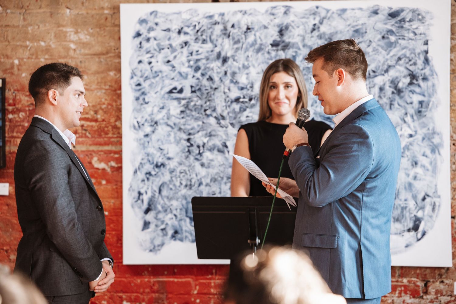 Wedding ceremony with officiant reading vows as couple stands before abstract art in brick venue.