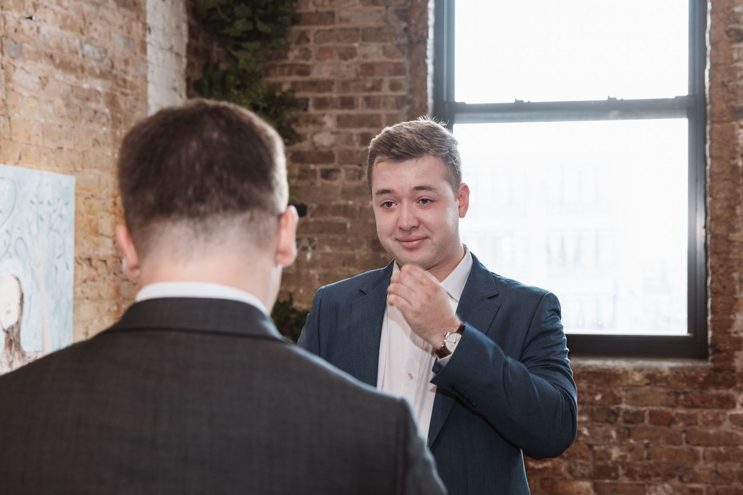 Two men in suits converse near a bright window in a brick-walled room.