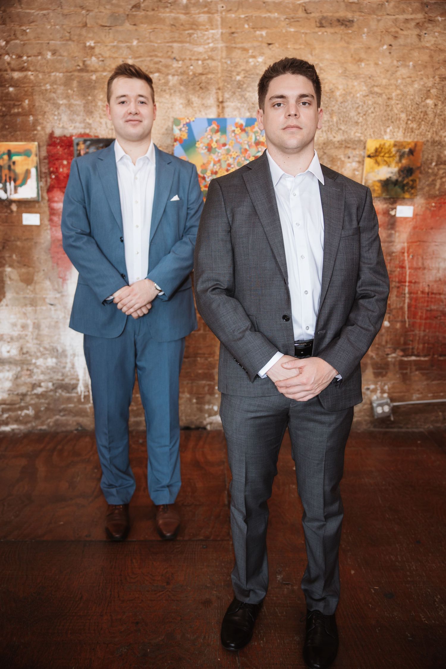 Two well-dressed young men in blue and gray suits standing in a rustic brick art gallery.