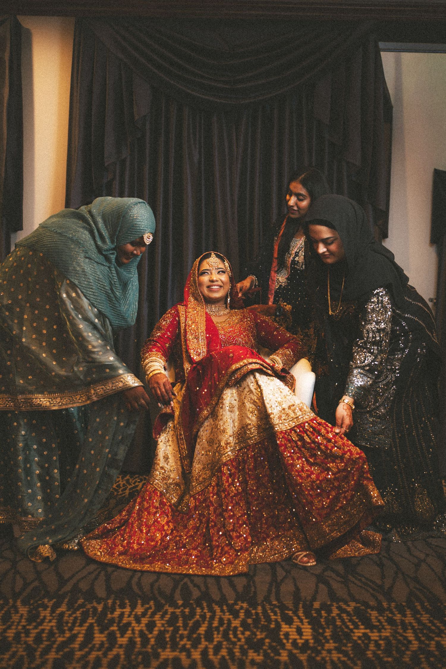 Smiling South Asian bride in red bridal lehenga is assisted by attendants in a room.
