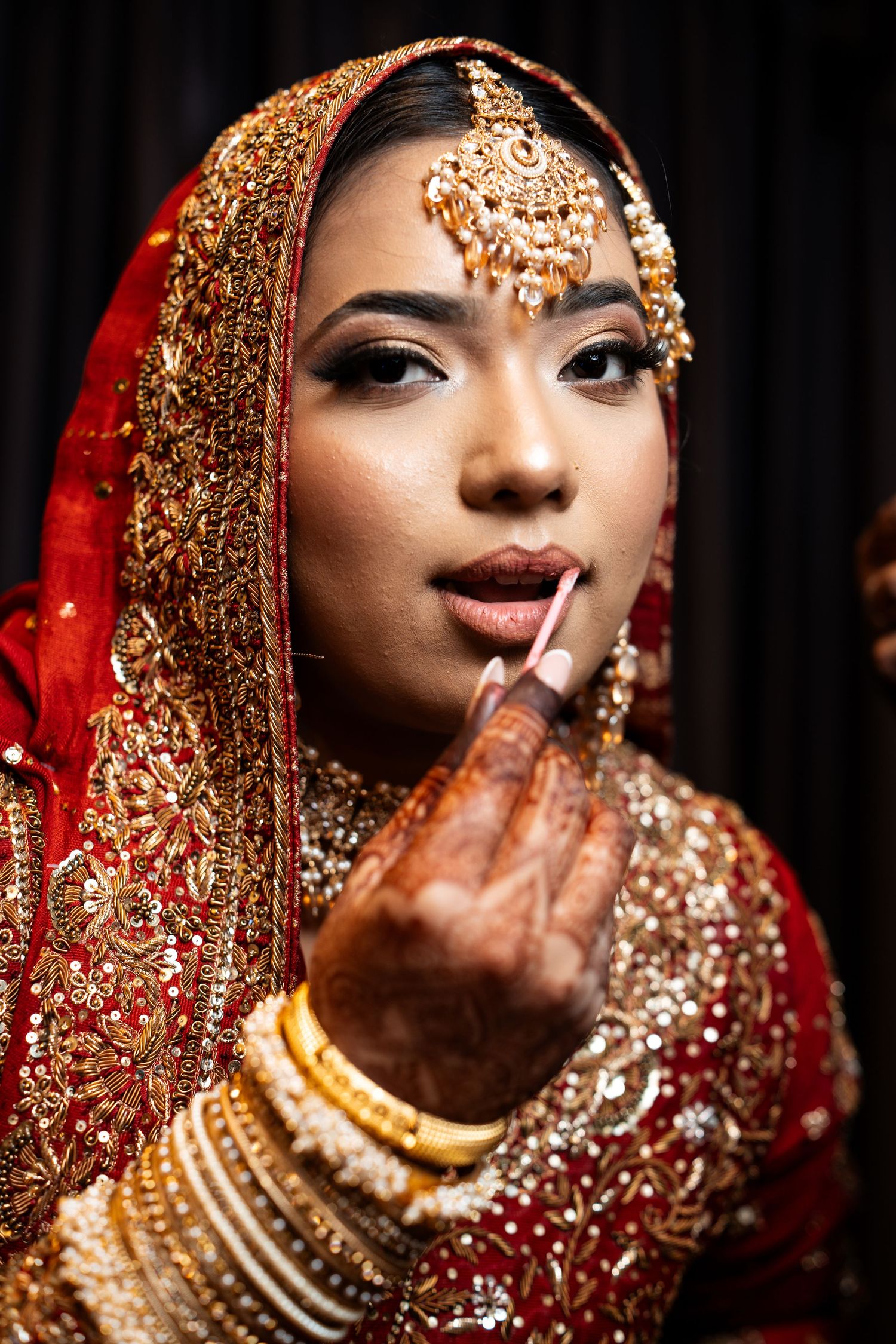A beautiful Indian bride in traditional red and gold lehenga has her makeup done before her wedding ceremony.