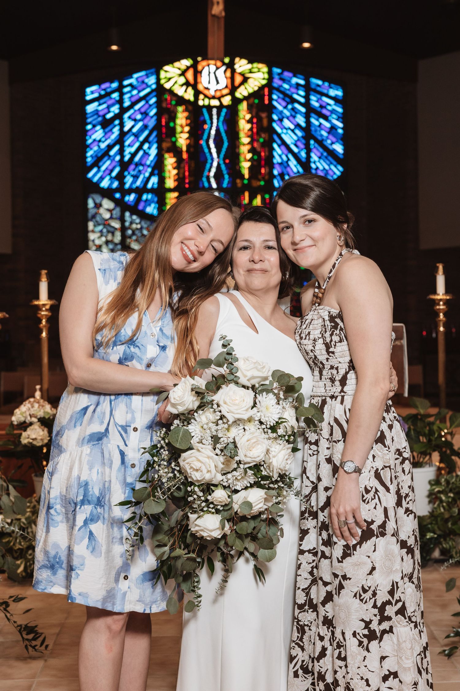 Bride holding white bouquet poses with two women in floral dresses before colorful stained glass church window.