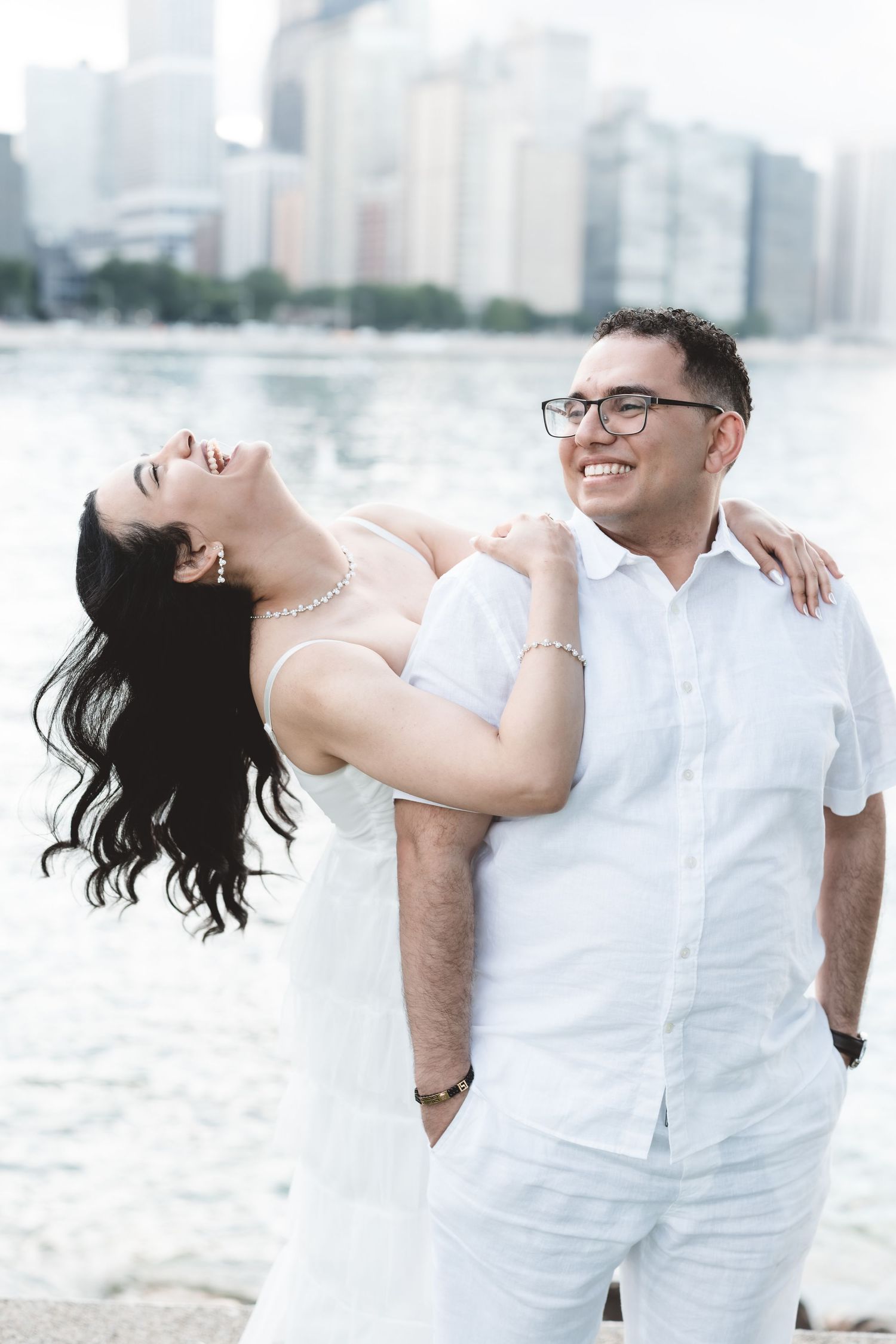 A joyful couple in matching white outfits laughing together by a city waterfront.