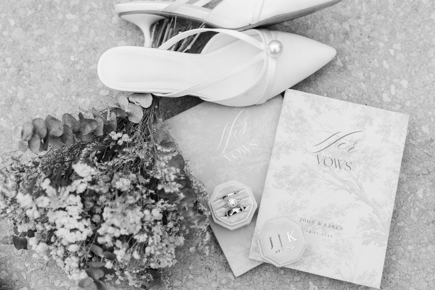Black and white flat lay of bridal details including heels, floral bouquet, vow books, and wedding rings on textured surface.