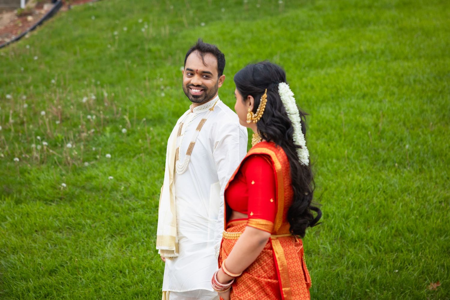 Indian wedding couple in traditional attire on green lawn, groom in white kurta smiling at bride in red saree.