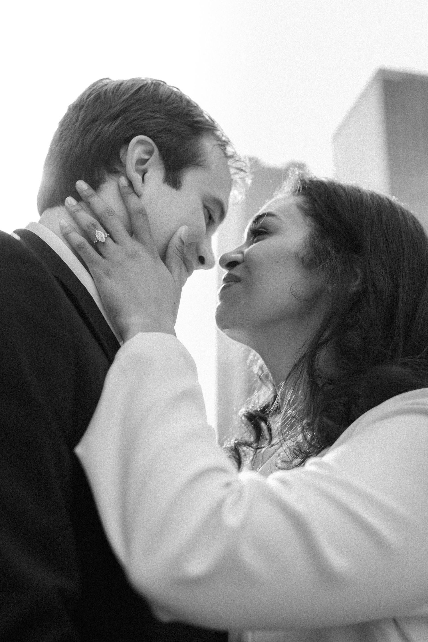 Black and white photo of couple sharing a tender kiss with city buildings behind them.