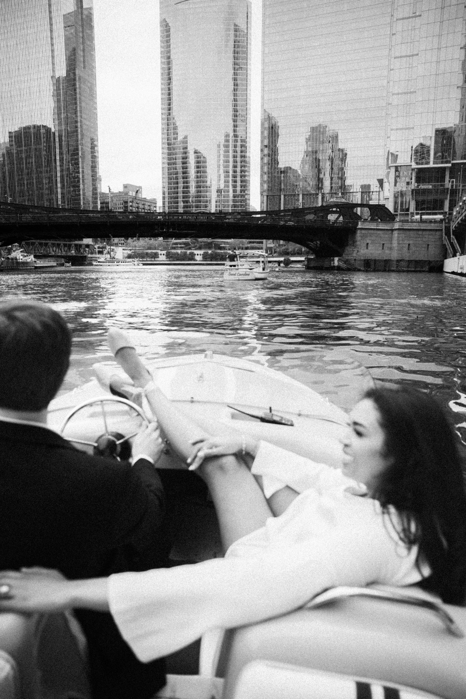 Couple holding hands on a boat with Chicago River skyline and bridge in black and white.
