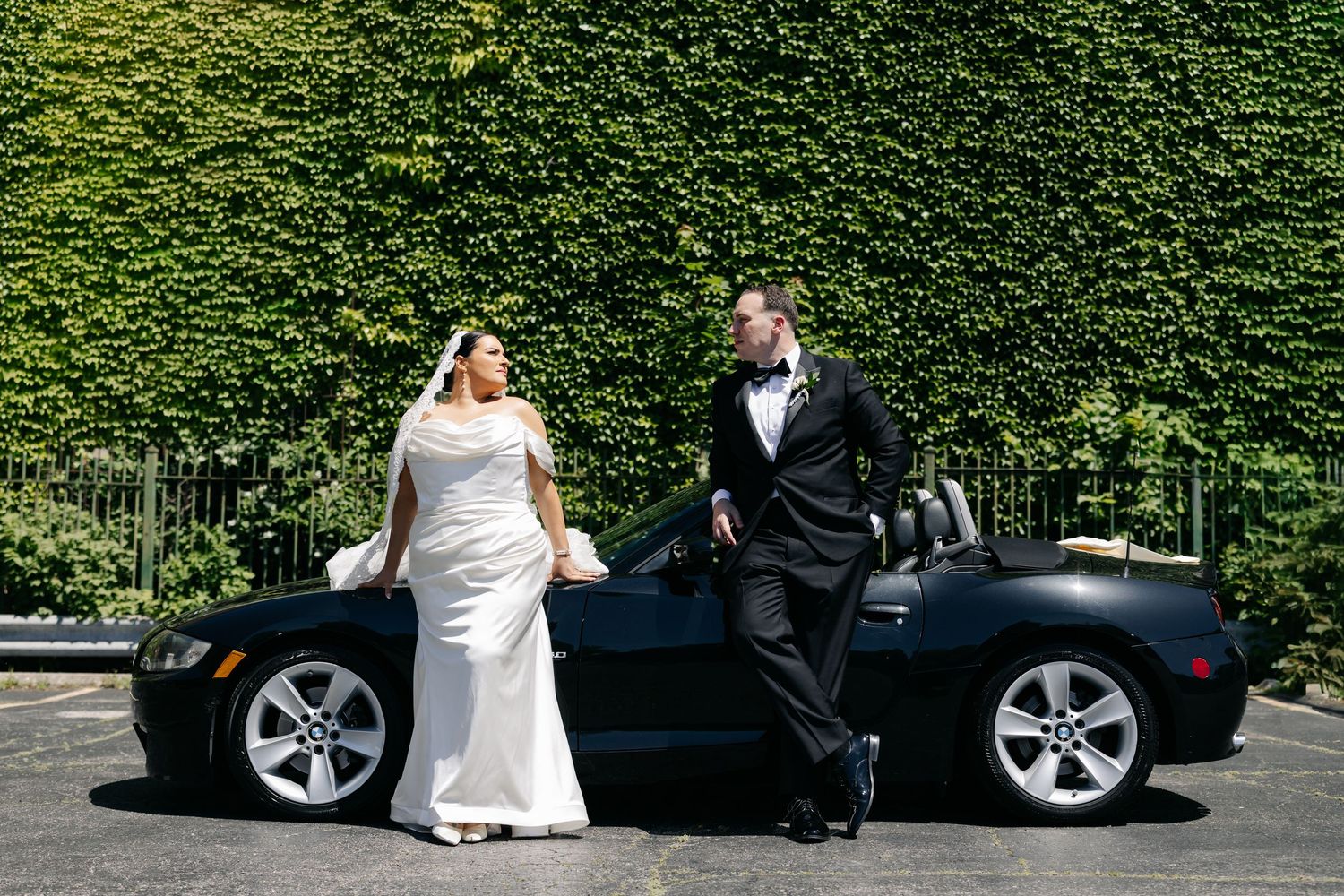 Bride and groom pose beside black convertible sports car outdoors.