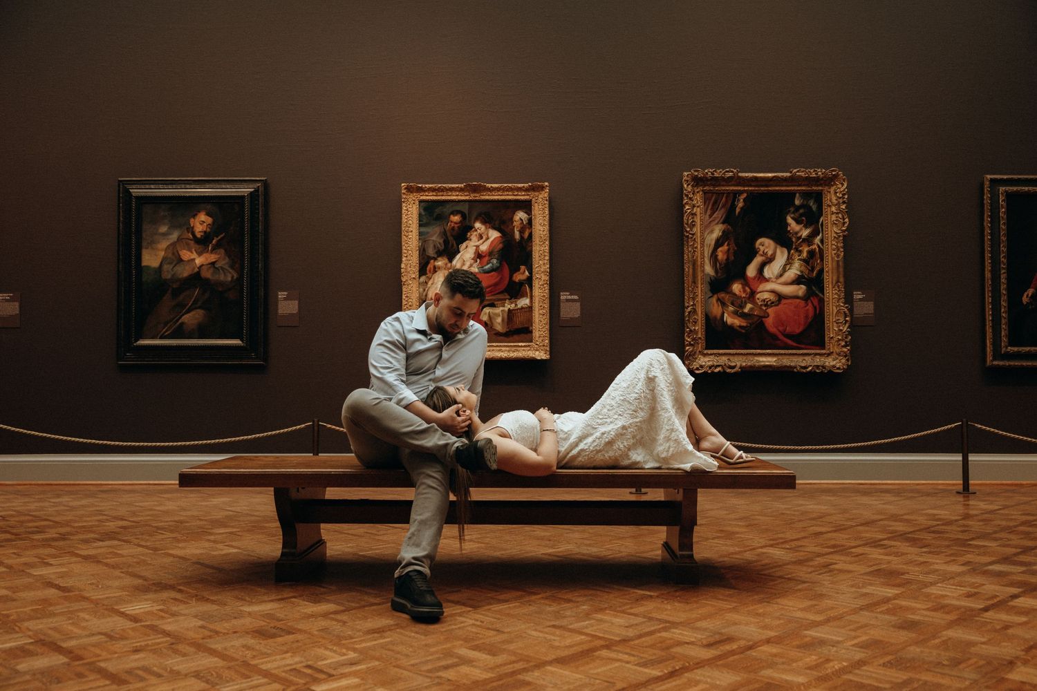 Romantic couple in formal wear relaxing on a museum bench surrounded by classical paintings in an art gallery.