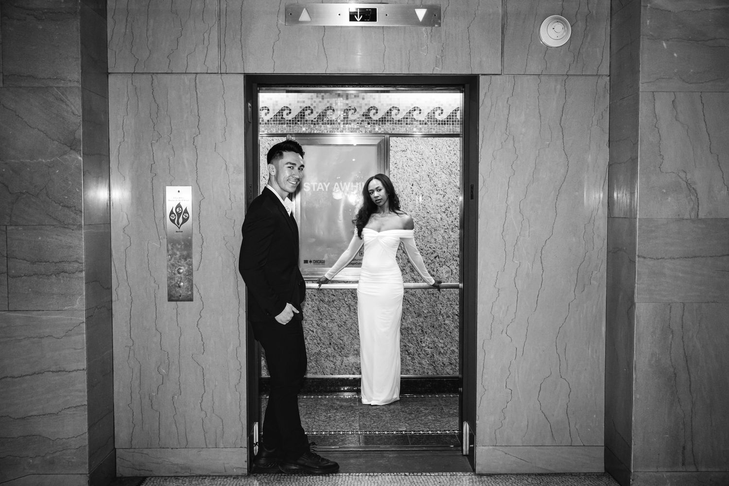 Black and white photo of a stylish couple posing inside an elegant elevator on their wedding day.