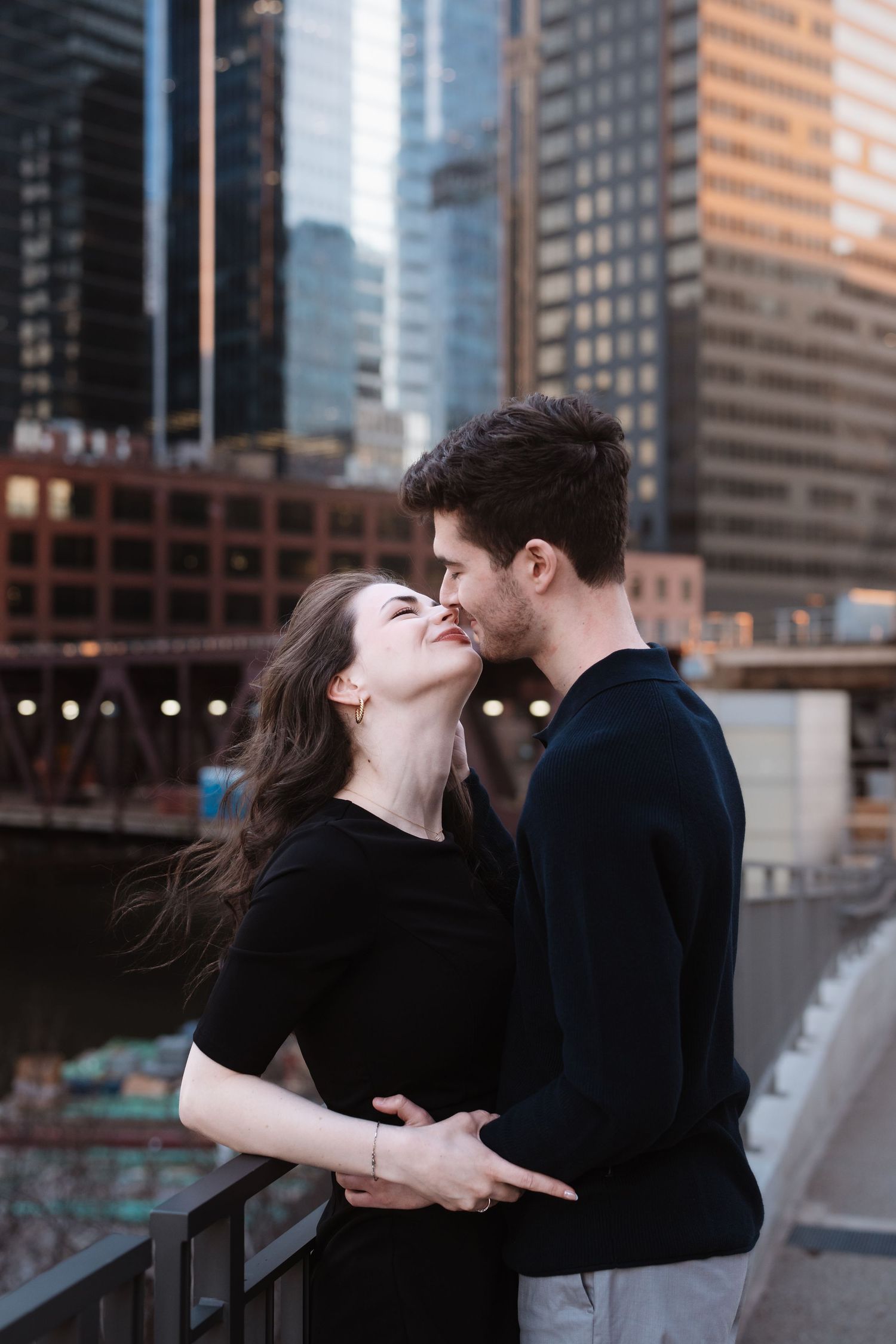 A happy couple almost kissing while embracing on a bridge with Chicago skyscrapers behind them.