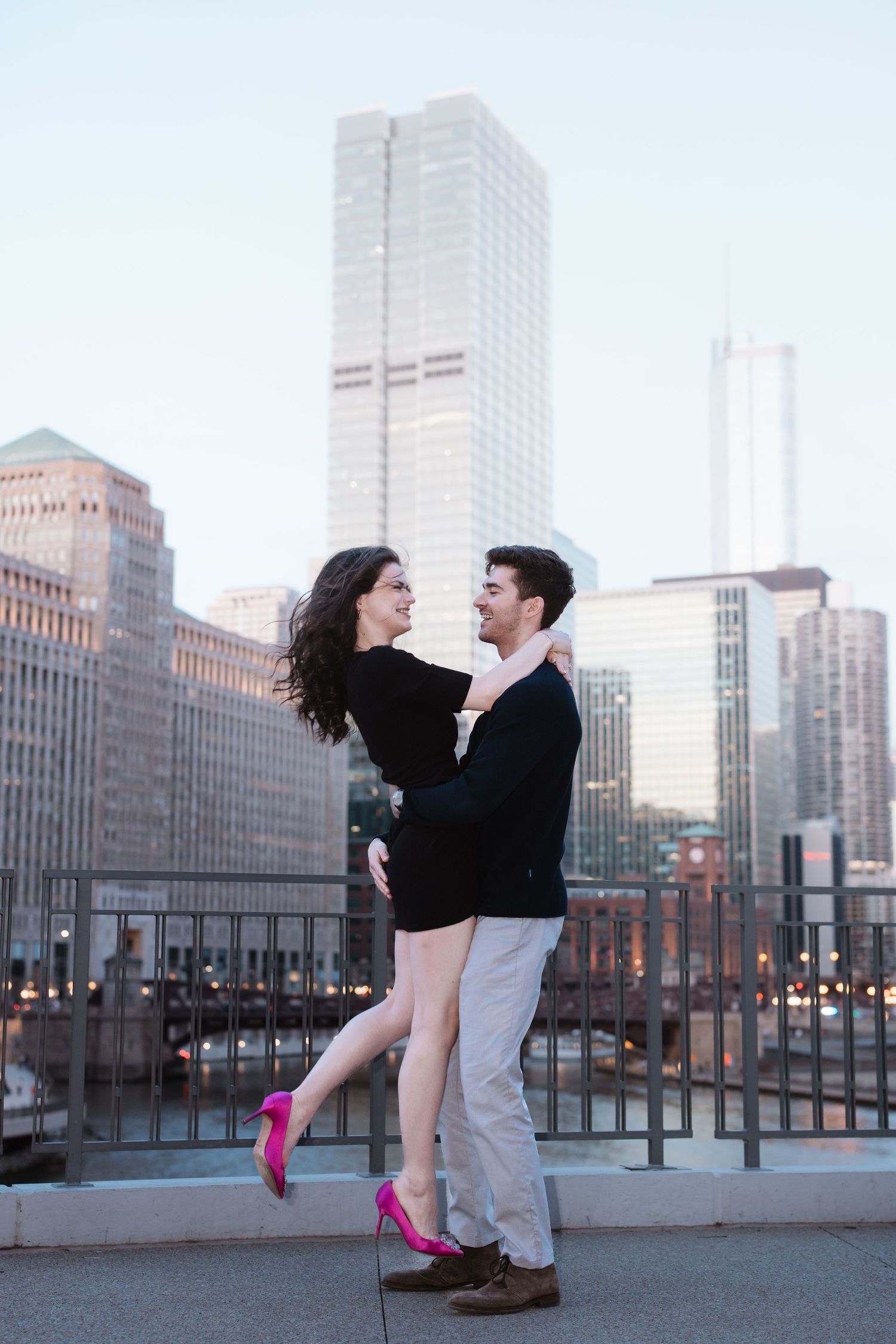 Man lifting smiling woman in pink heels with Chicago skyline and river in background.
