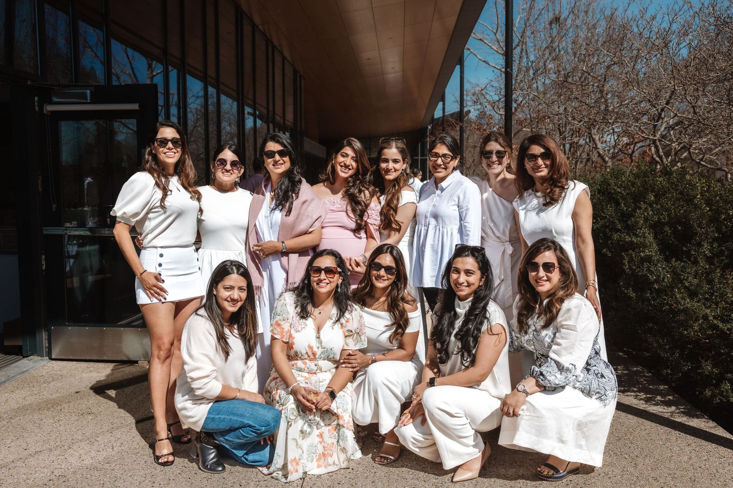 Group of women in white outfits posing together outdoors at a sunny spring baby shower celebration.