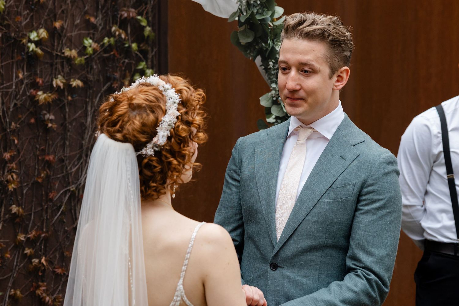 Groom in gray suit smiling at bride in wedding veil and hair accessory during outdoor ceremony.