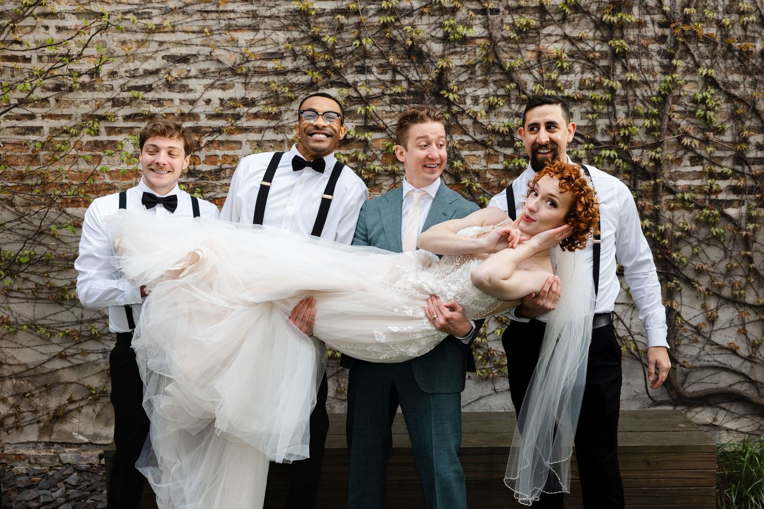 Groom in teal suit stands smiling as groomsmen in suspenders carry laughing red-haired bride in white wedding gown.