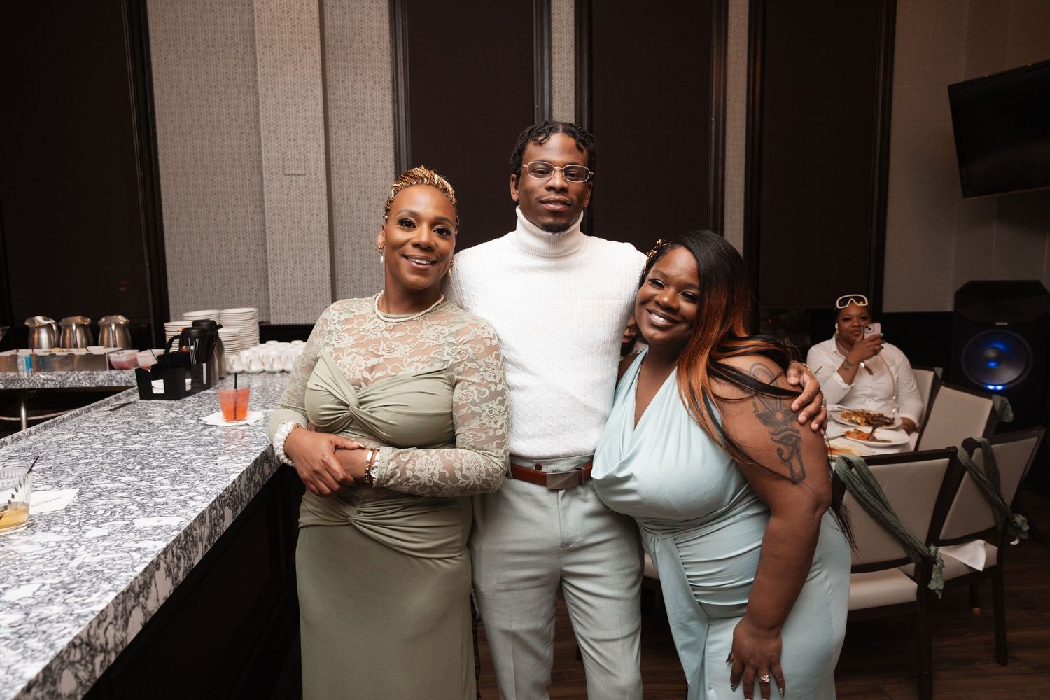Three guests pose together smiling near bar at wedding reception venue.