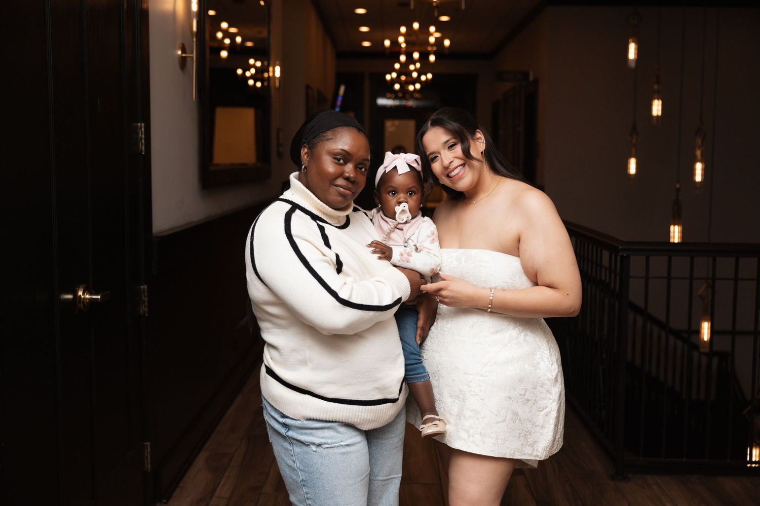 Woman in white sweater holds baby beside bride in white dress in warmly lit venue.