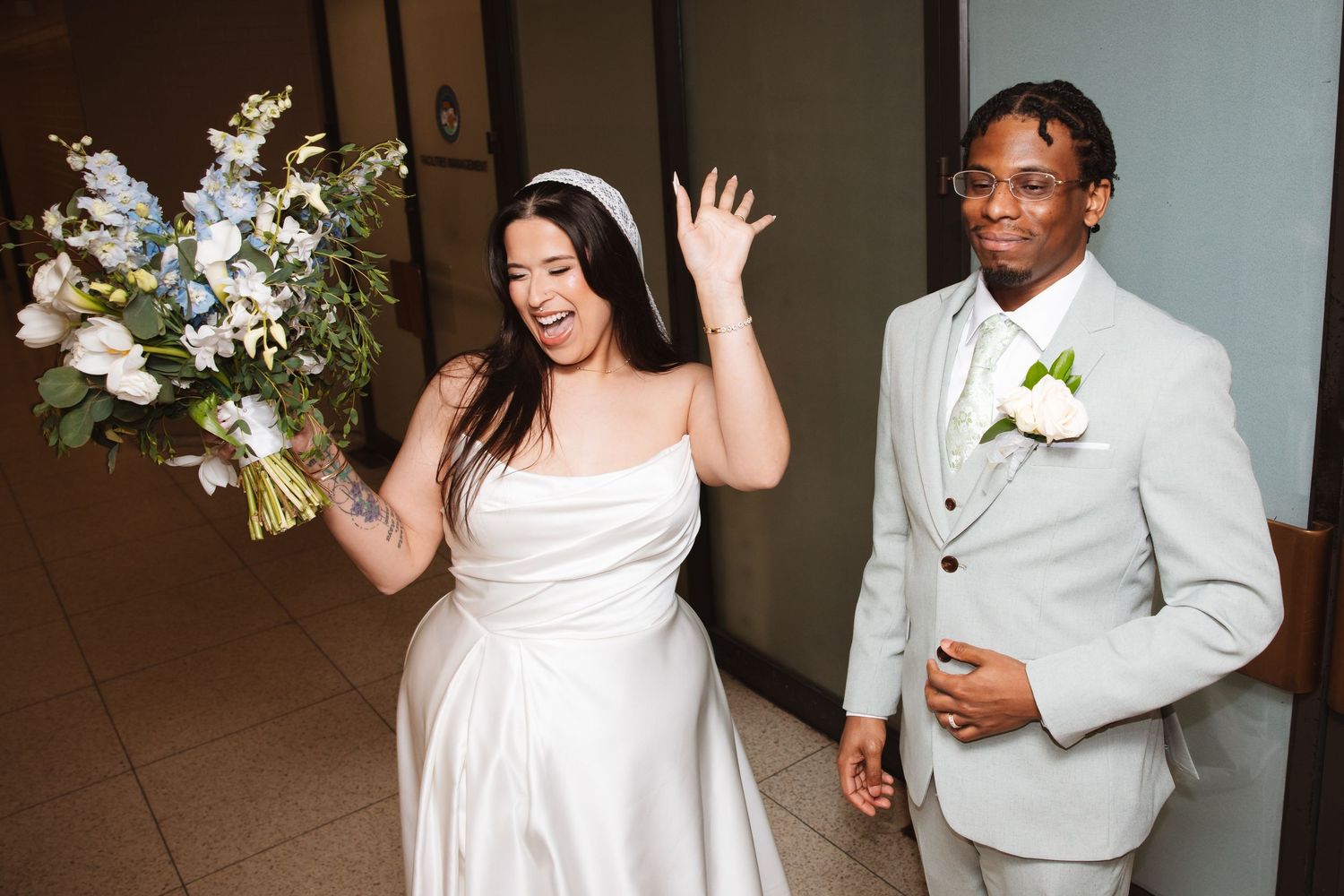 Joyful bride in white strapless gown waves while walking with groom in light gray suit holding bouquet.