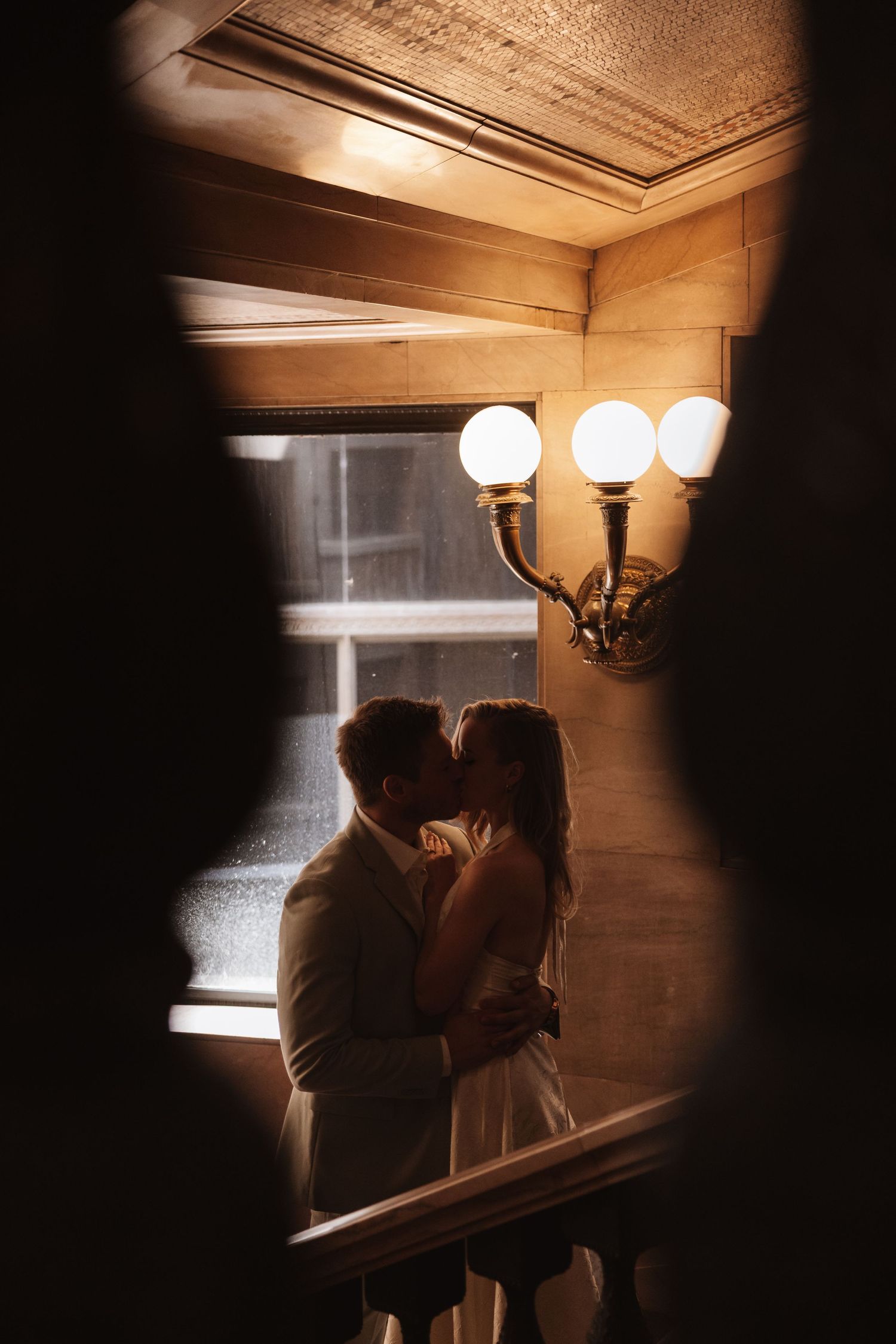 Bride and groom sharing a romantic kiss in dimly lit corridor with glowing wall light.