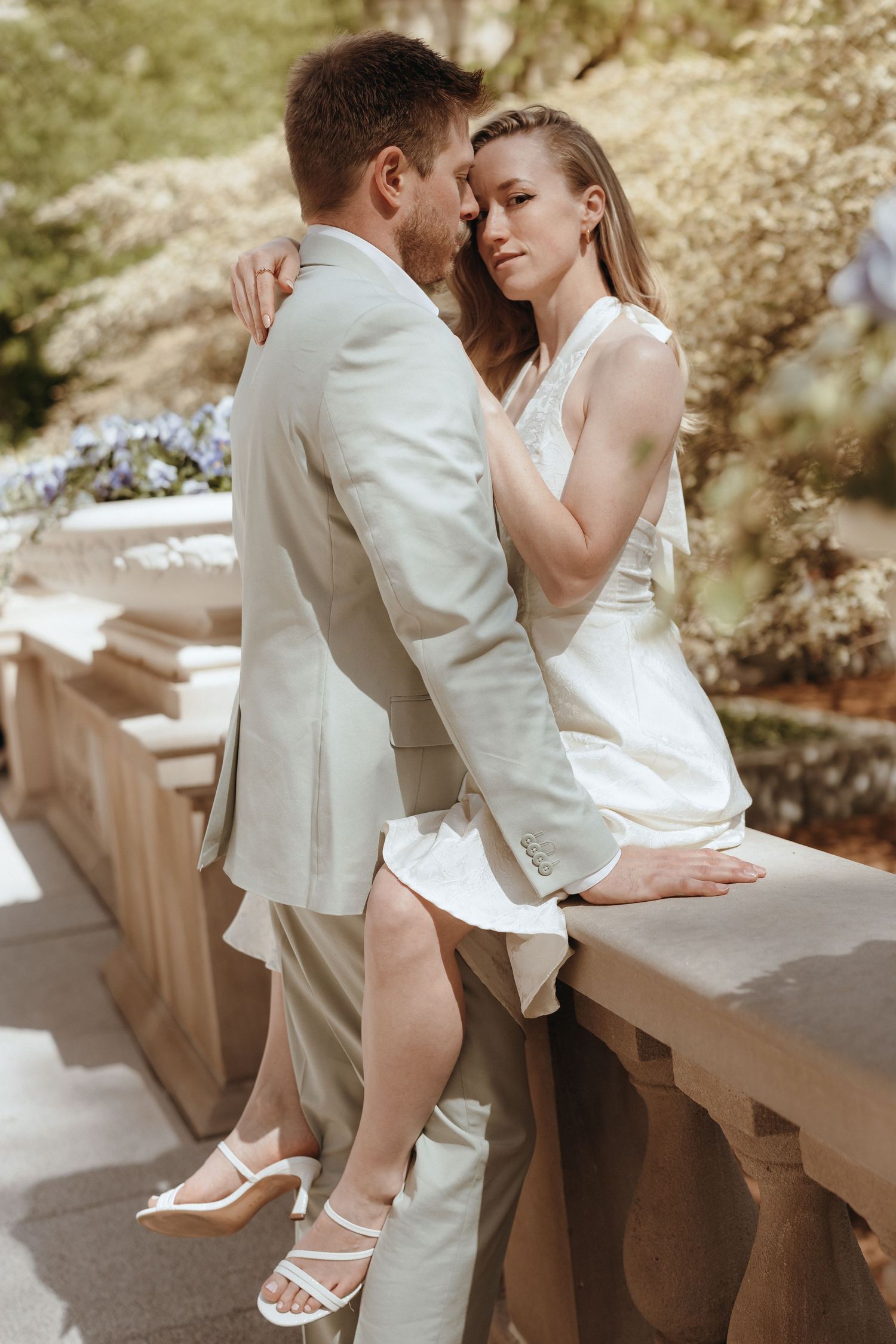 Bride in white mini dress sits on stone balustrade embracing groom in light suit in sunny garden.
