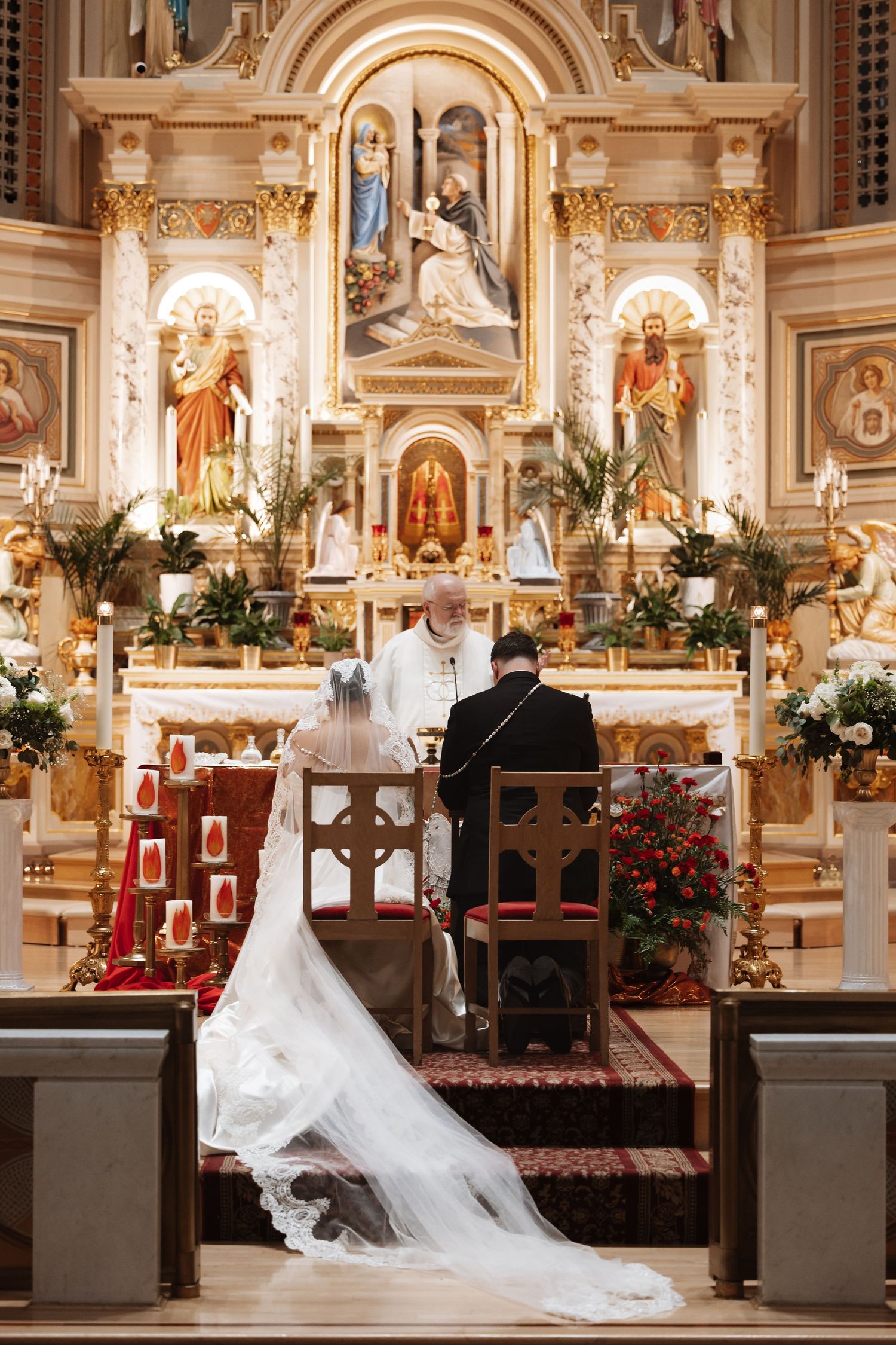 Bride and groom kneel before ornate golden altar during Catholic wedding ceremony with priest officiating.
