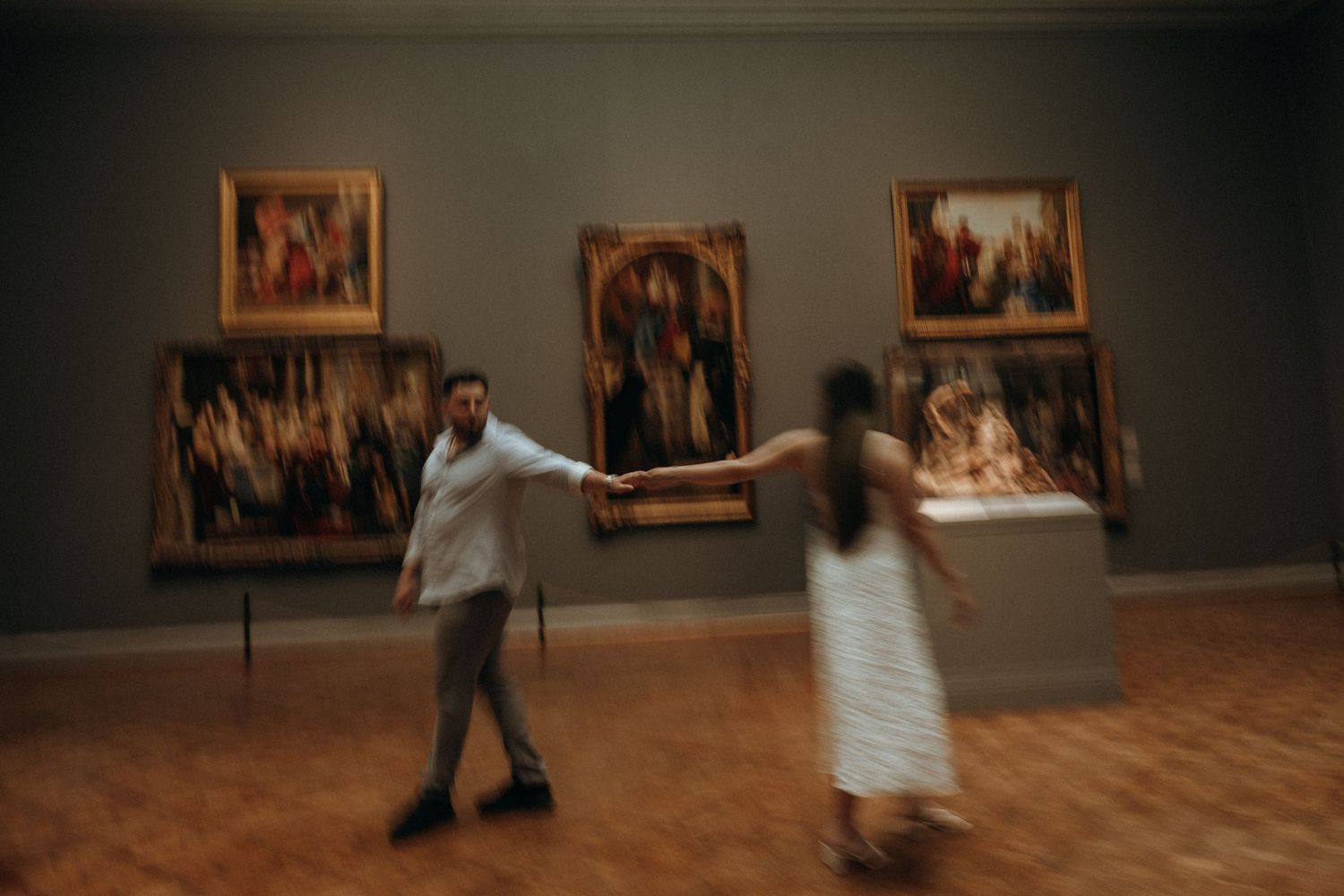 Couple dancing playfully with motion blur effect in front of colorful paintings in an art museum.