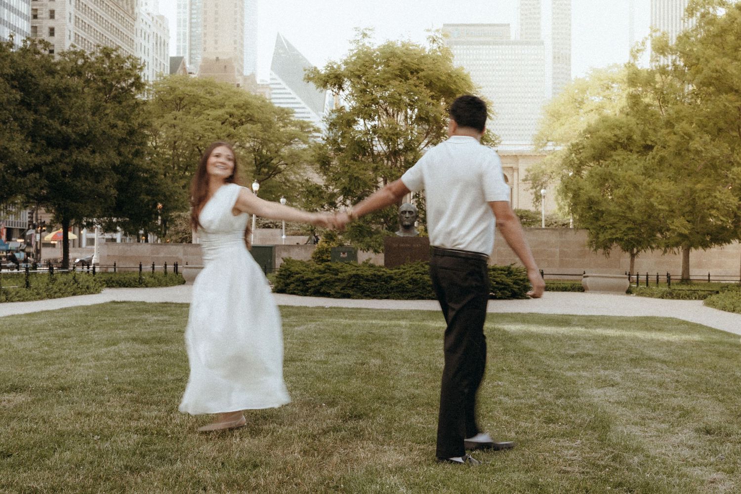 Engaged couple dancing on green lawn in urban park, woman in white dress, man in white shirt.