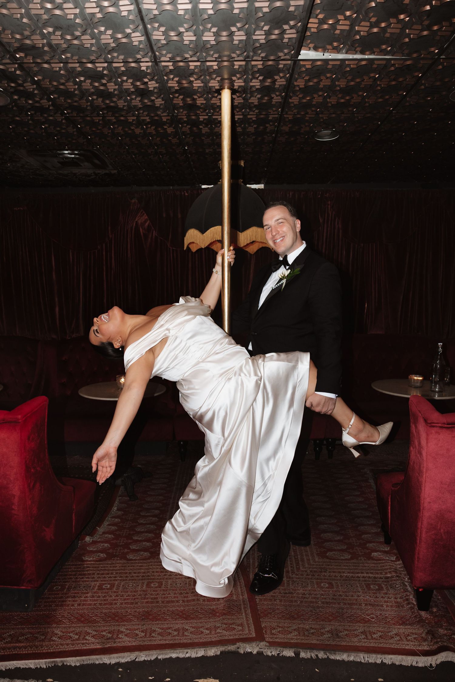 Bride playfully poses on gold pole while groom smiles in dark red velvet reception venue.