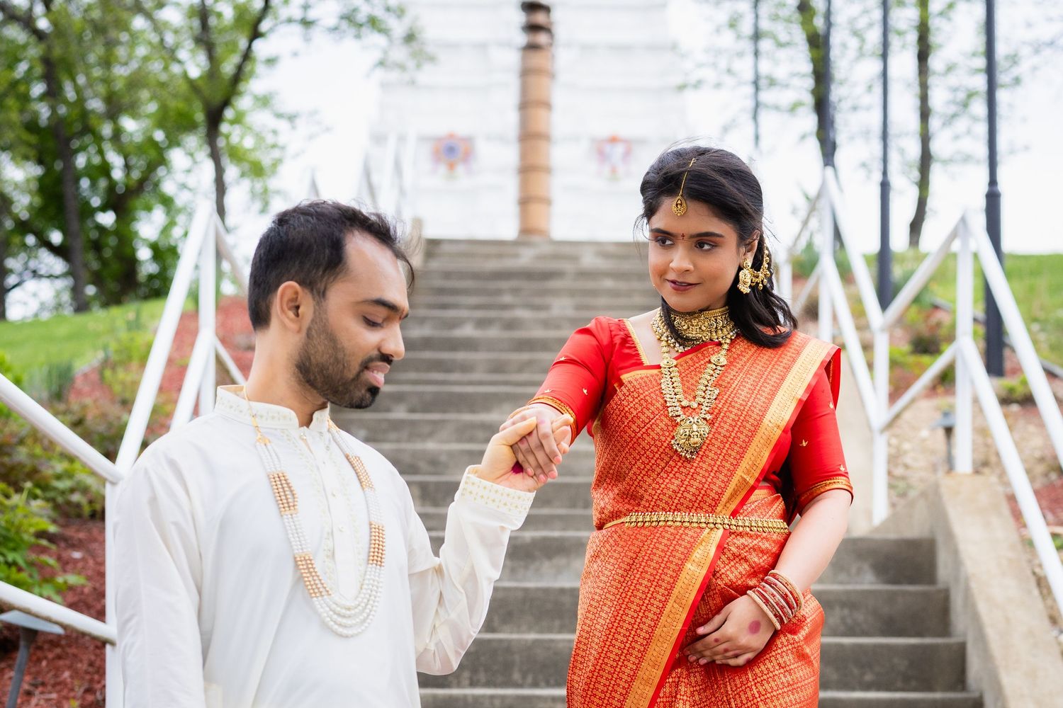 Indian couple in traditional attire on temple steps, bride in red saree with gold jewelry, groom in white kurta holding hands.