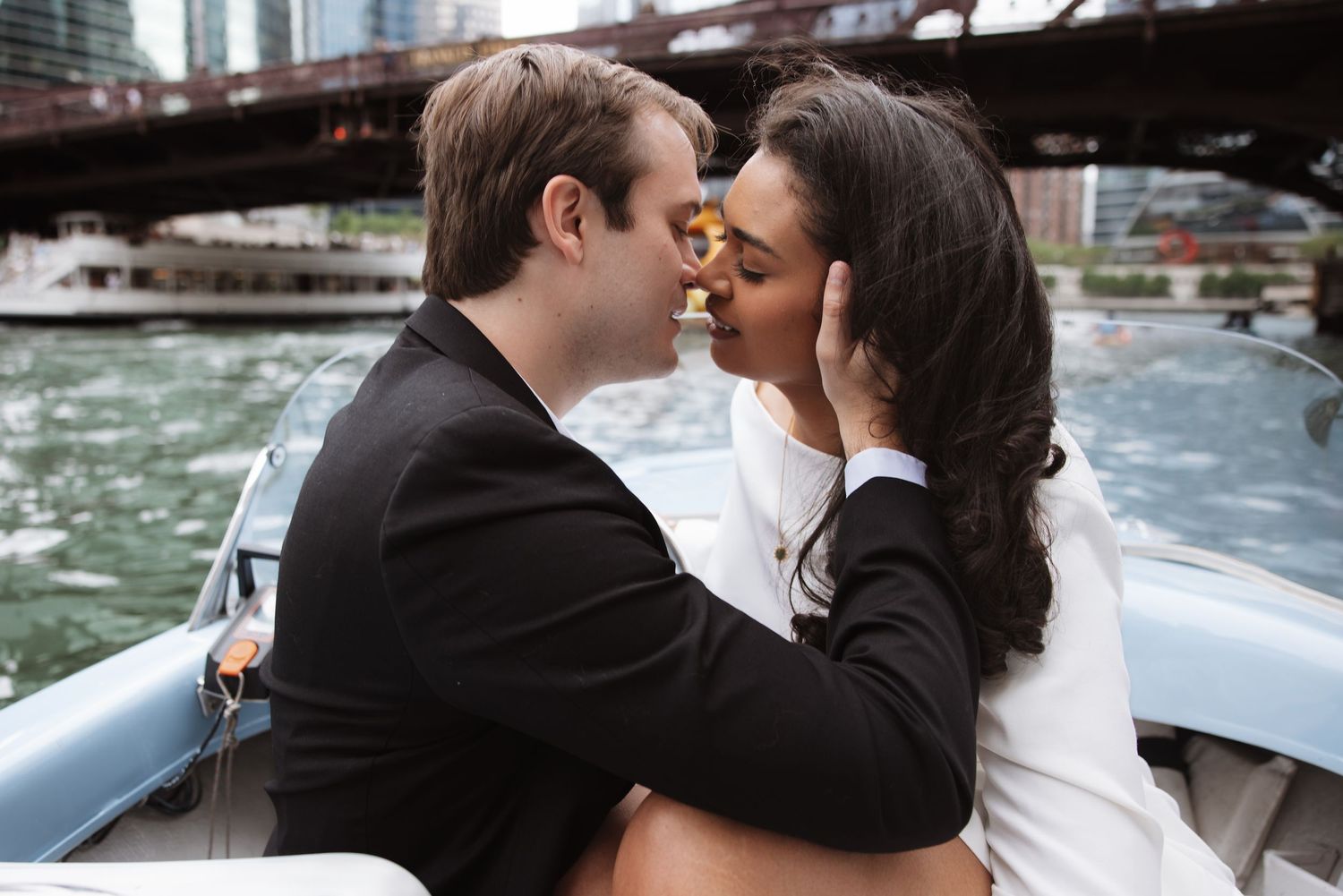 Couple sharing a romantic kiss on a boat with a city bridge and skyline in the background.