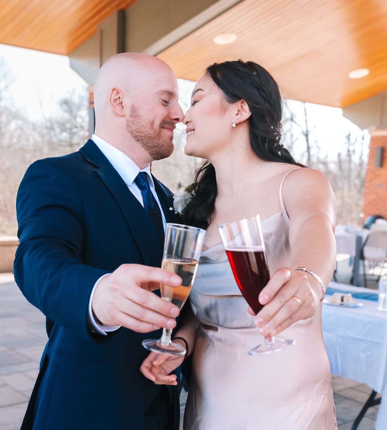 A newlywed couple clinks wine glasses and shares a kiss at their outdoor wedding reception.