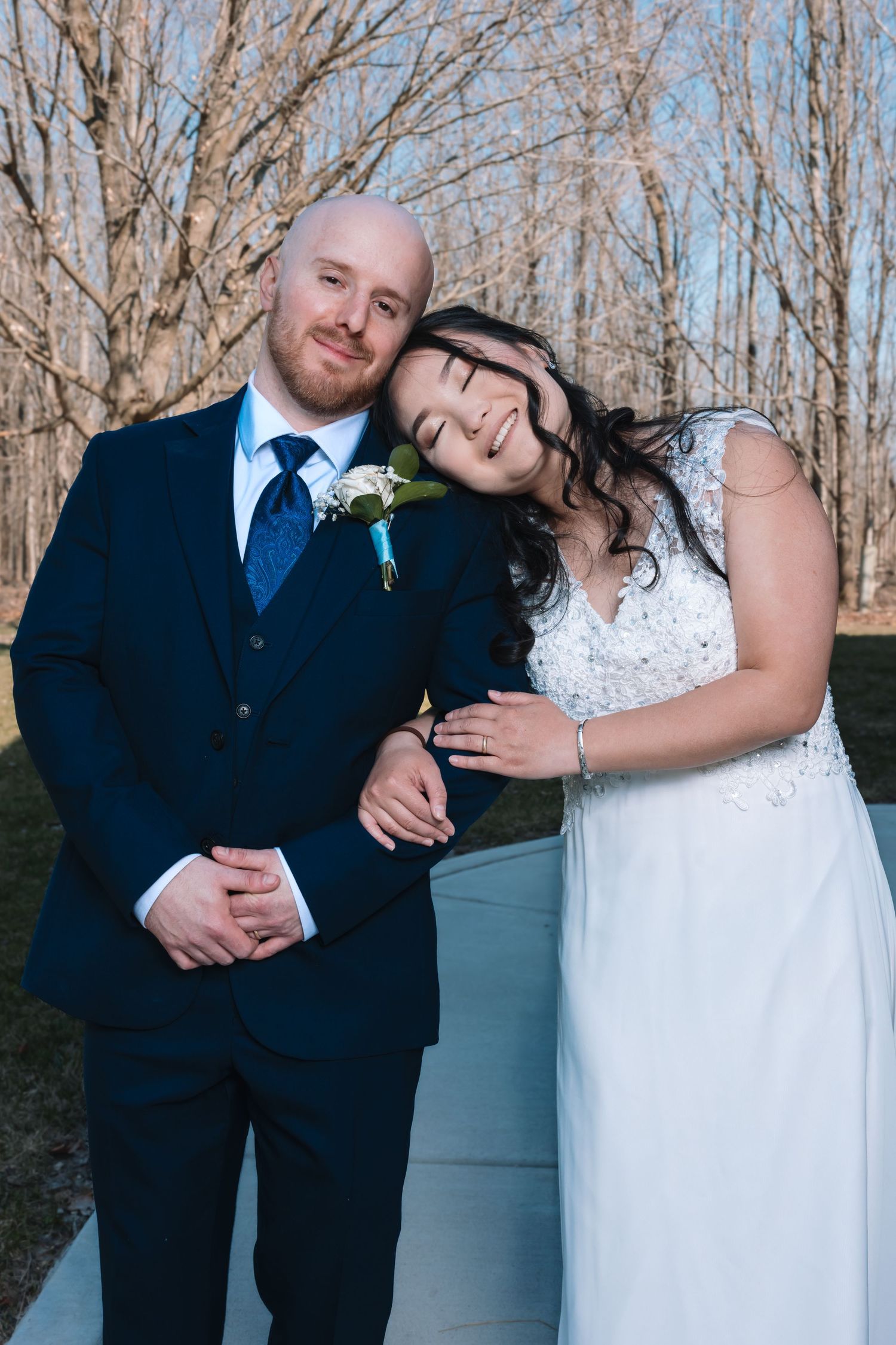 Bride leaning on groom's shoulder smiling in wedding attire outdoors.