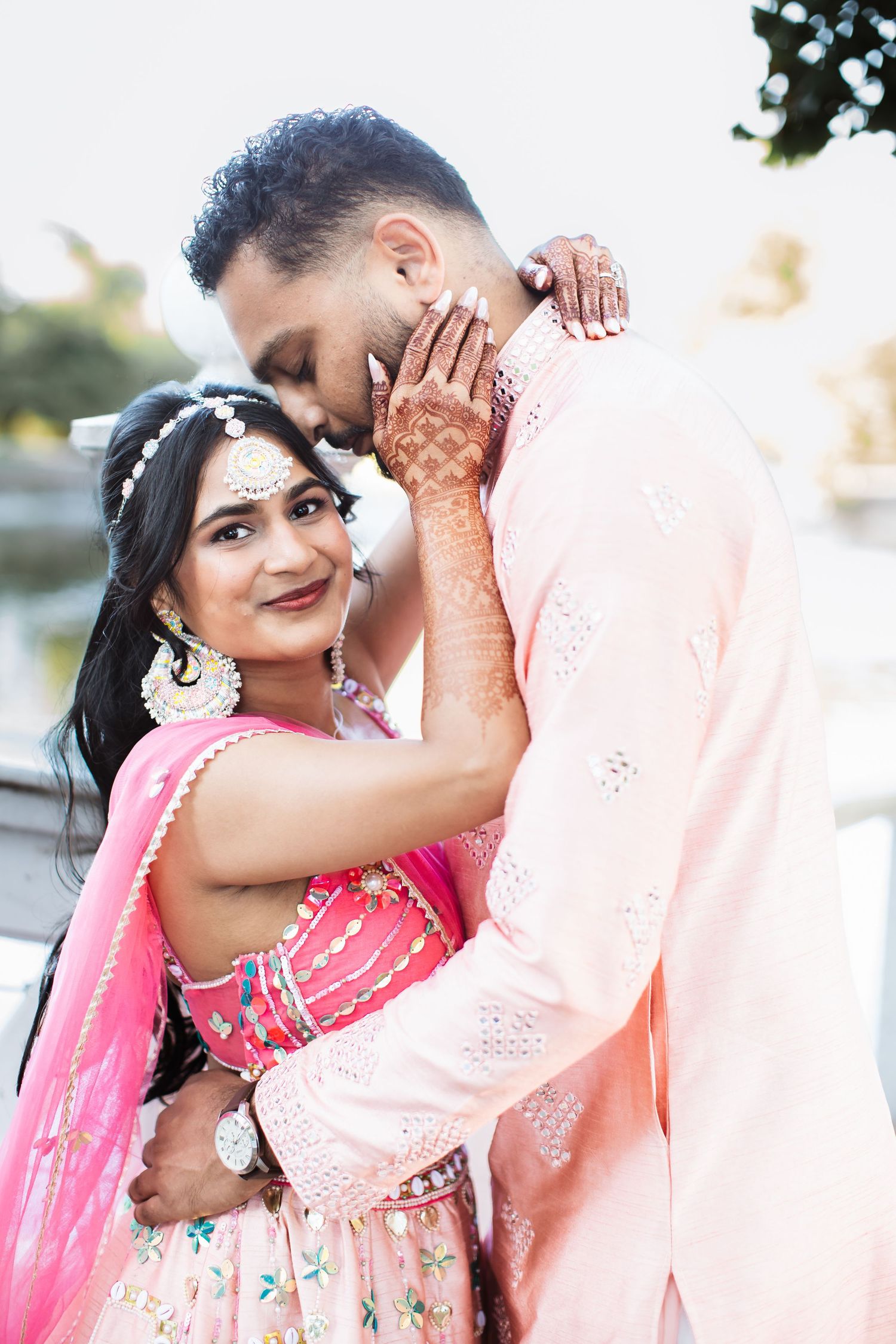 Indian couple in pink traditional attire embracing outdoors, groom kissing brides henna-adorned hand tenderly.