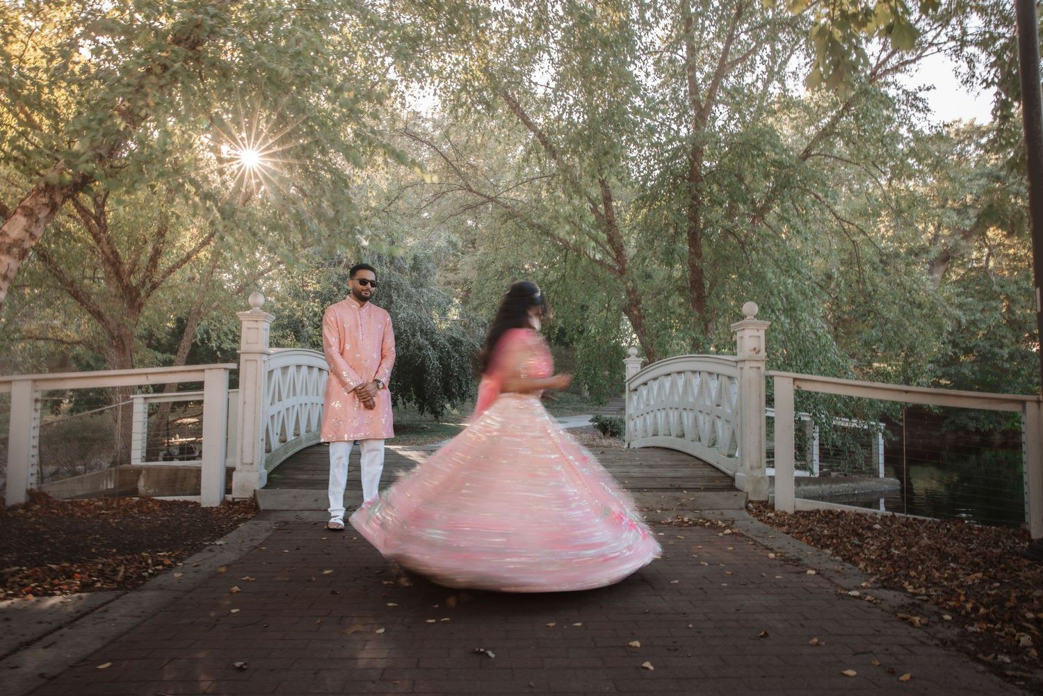 Indian couple in pink wedding attire with sunlight filtering through trees by bridge.