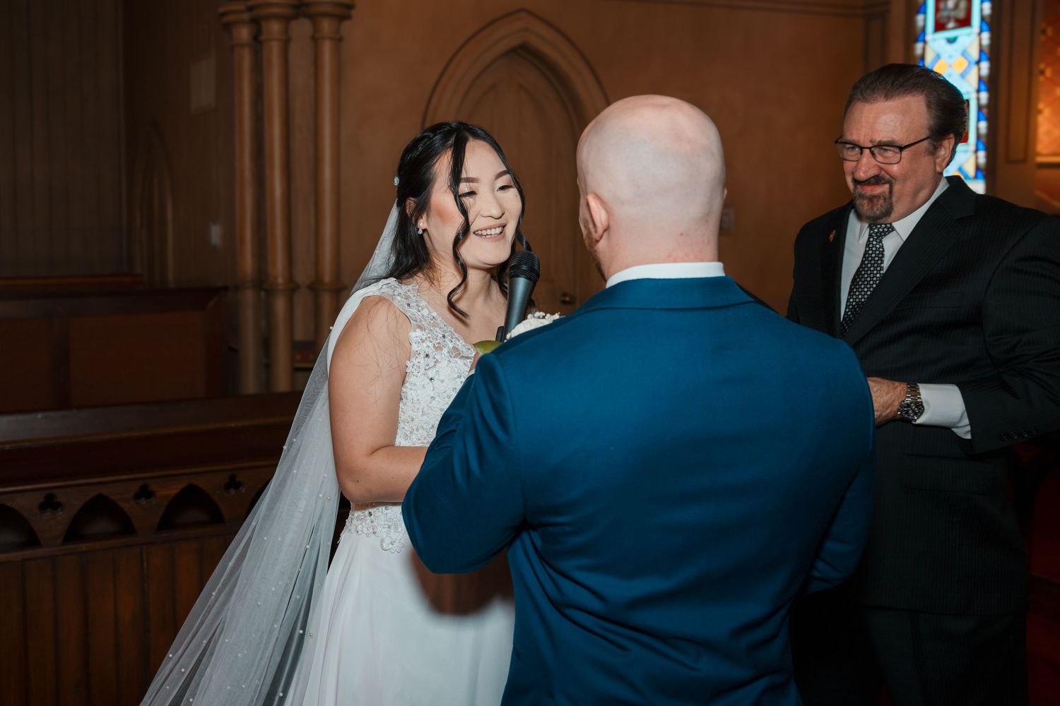 Bride laughing joyfully at groom during wedding ceremony with officiant beside them in church.