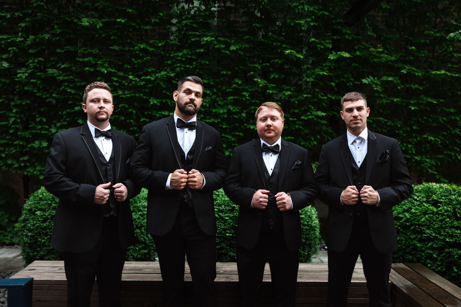 Four groomsmen in black tuxedos with bow ties adjusting their jackets outdoors against a lush green ivy wall.