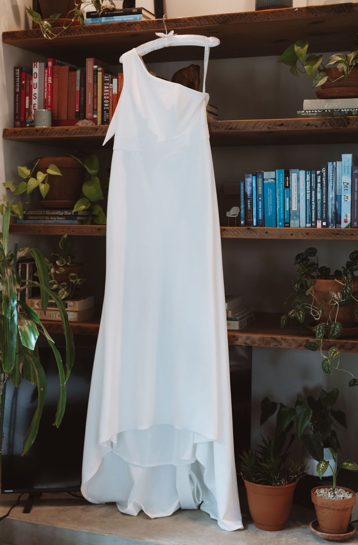 Simple white one-shoulder wedding gown hanging on a hanger in front of a bookshelf with plants.