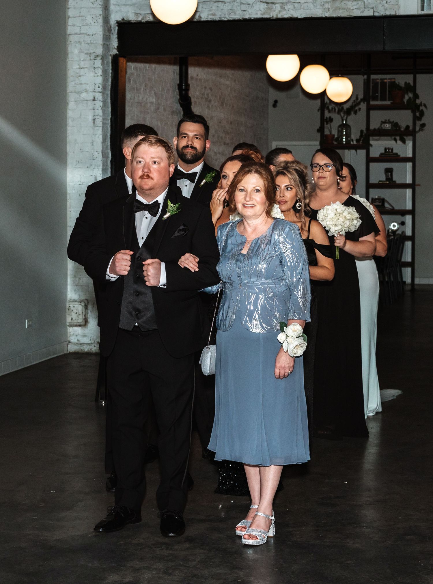 Man in tuxedo escorting woman in blue gown down aisle with wedding party behind.