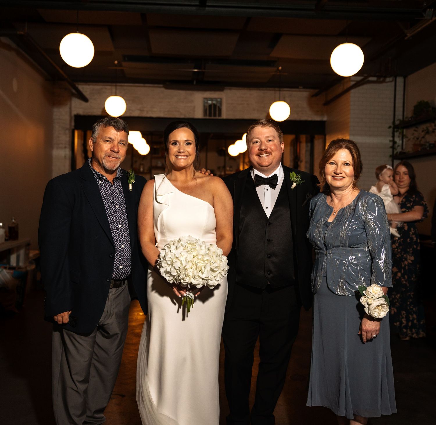 Bride in one-shoulder gown holds white bouquet posing with groom and two family members at reception.
