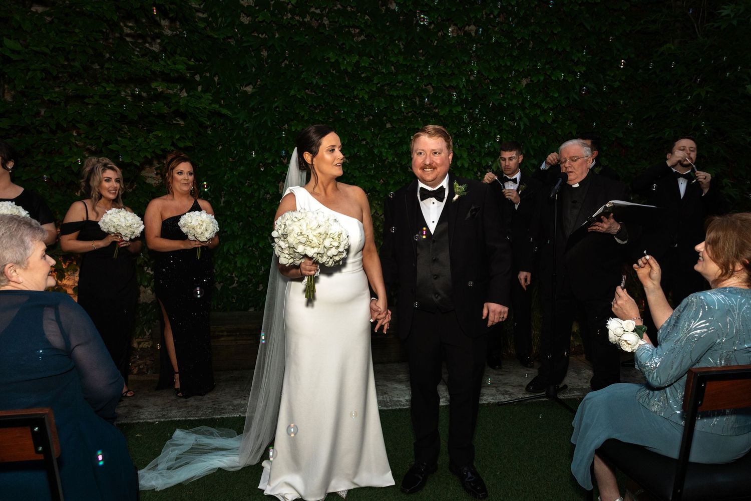 Bride and groom smile joyfully at outdoor wedding ceremony with bridesmaids in black dresses and green ivy backdrop.